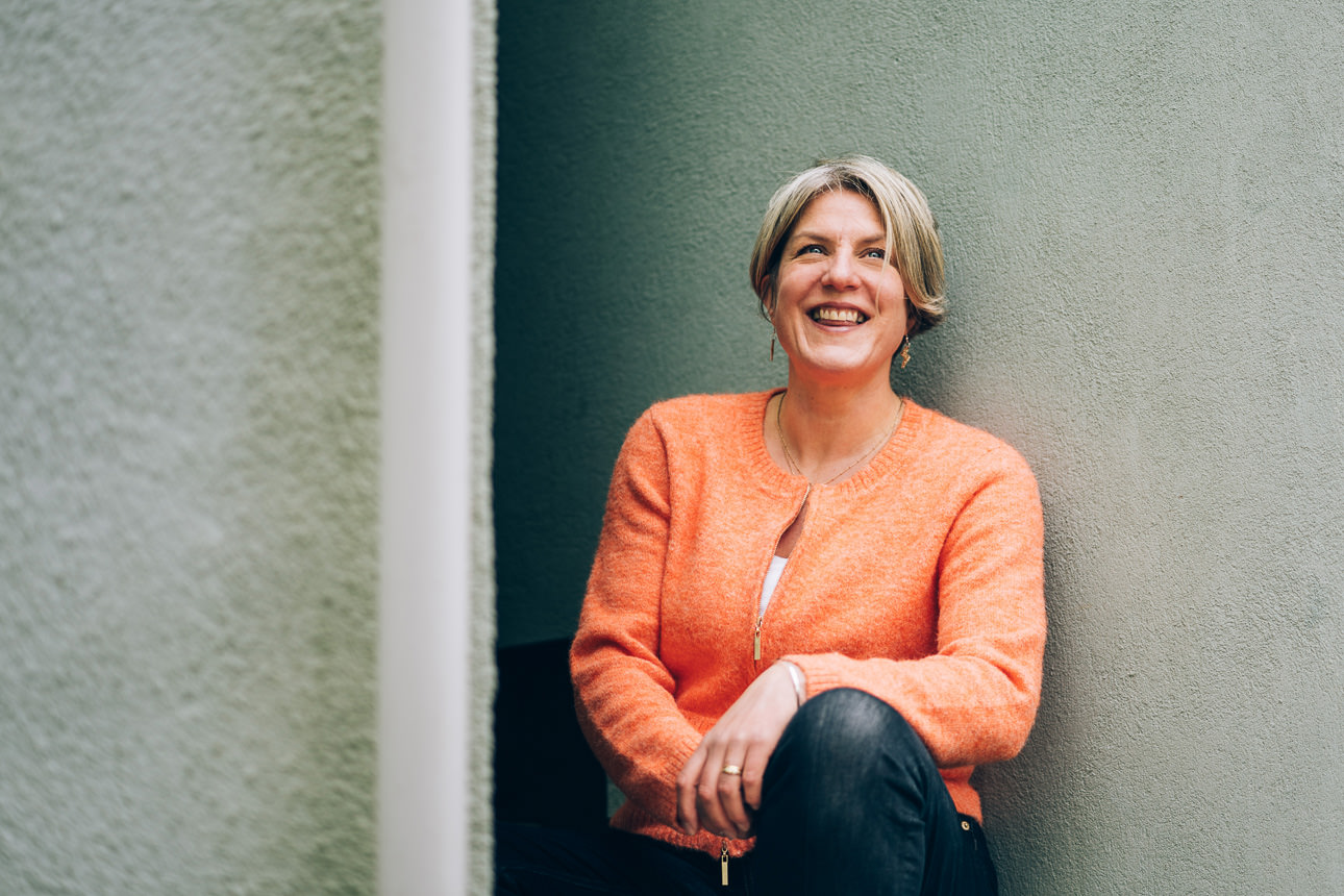Devon wedding and family photographer, Helen Lisk, in an orange cardigan sitting in front of a green wall