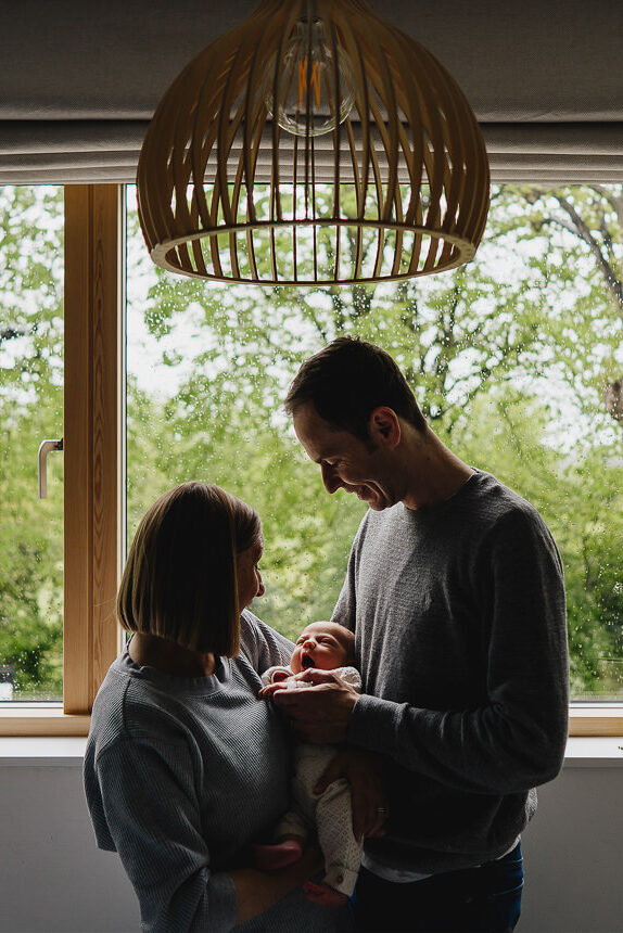 A couple of parents holding a newborn baby at their home, standing inside by a large window with trees outside
