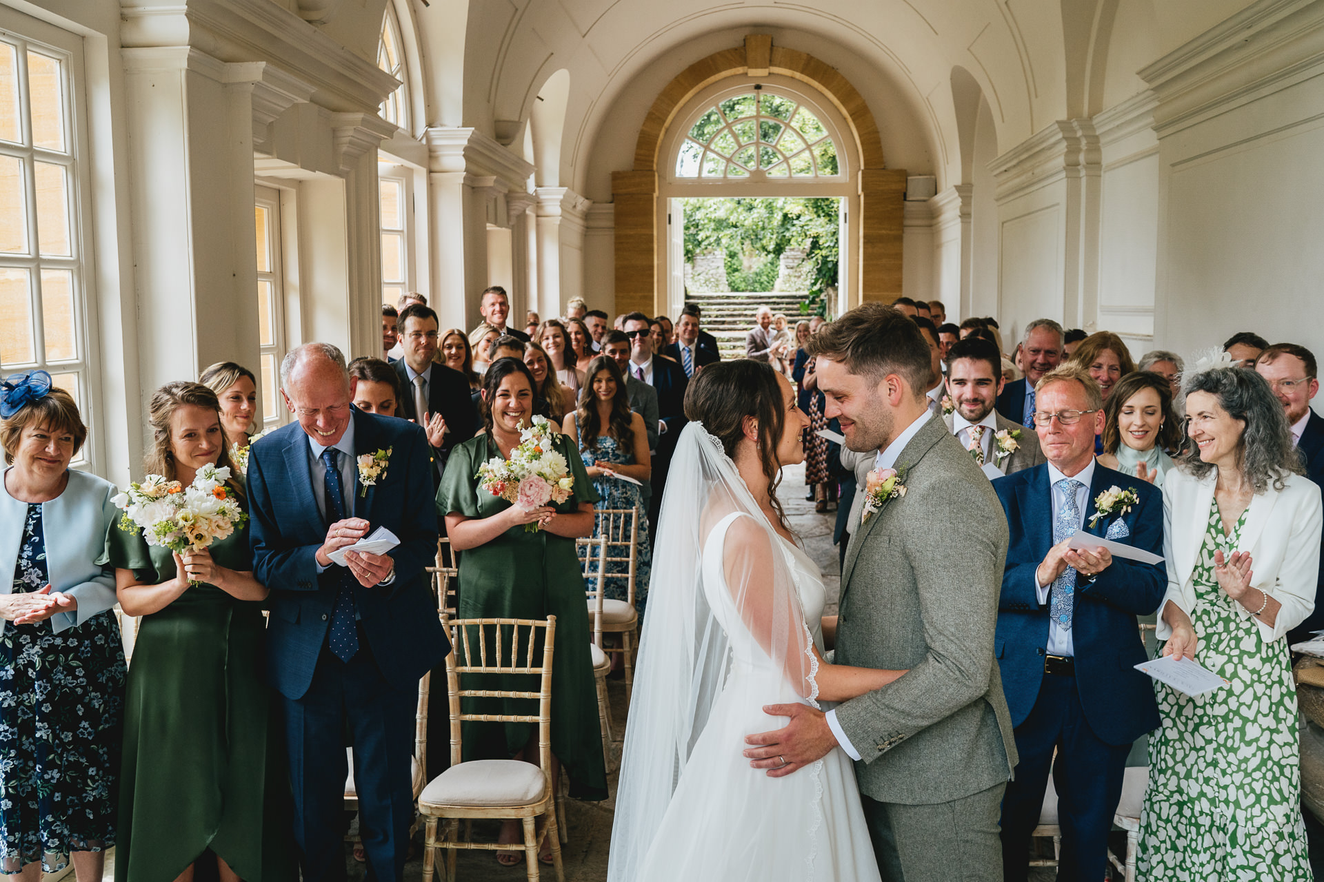 A bride and groom during their wedding ceremony in the Orangerie at Hestercombe Gardens in Somerset. The couple are cuddling and smiling at each other and the weddings guests are clapping behind them.