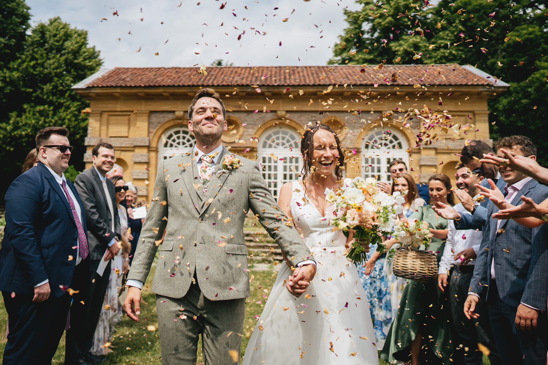 A bride and groom walking through confetti with an orangery behind them