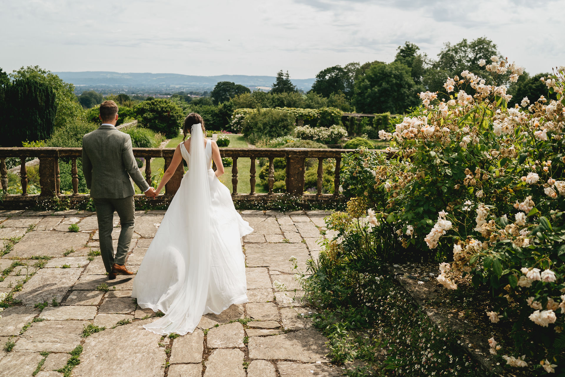 A groom and bride walking across a stone terrace with beautiful views of Somerset at Hestercombe Gardens