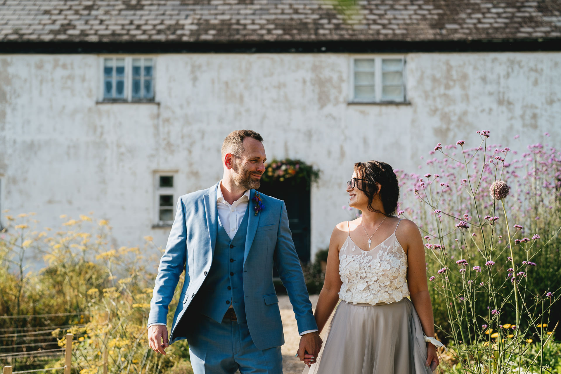 Bride and groom walking through the kitchen garden and smiling at each other with River Cottage behind them