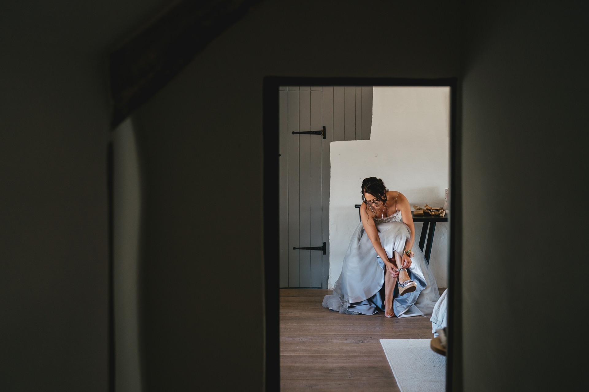 A bride in stylish separates putting on her shoes in the farmhouse at River Cottage