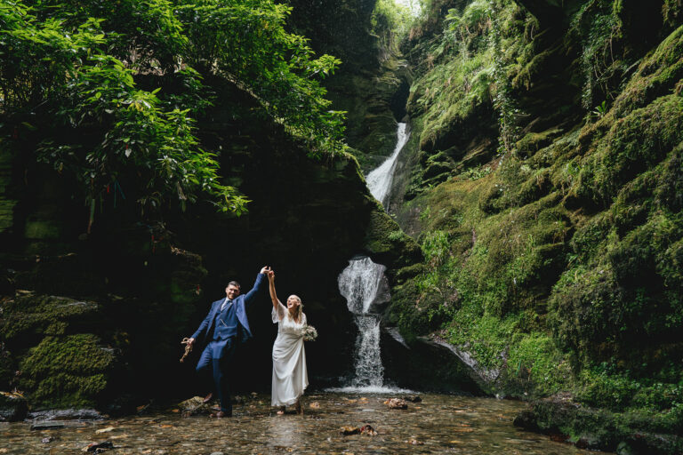 A bride and groom laughing and dancing in a pool by a waterfall during their St Nectan's Glen elopement wedding