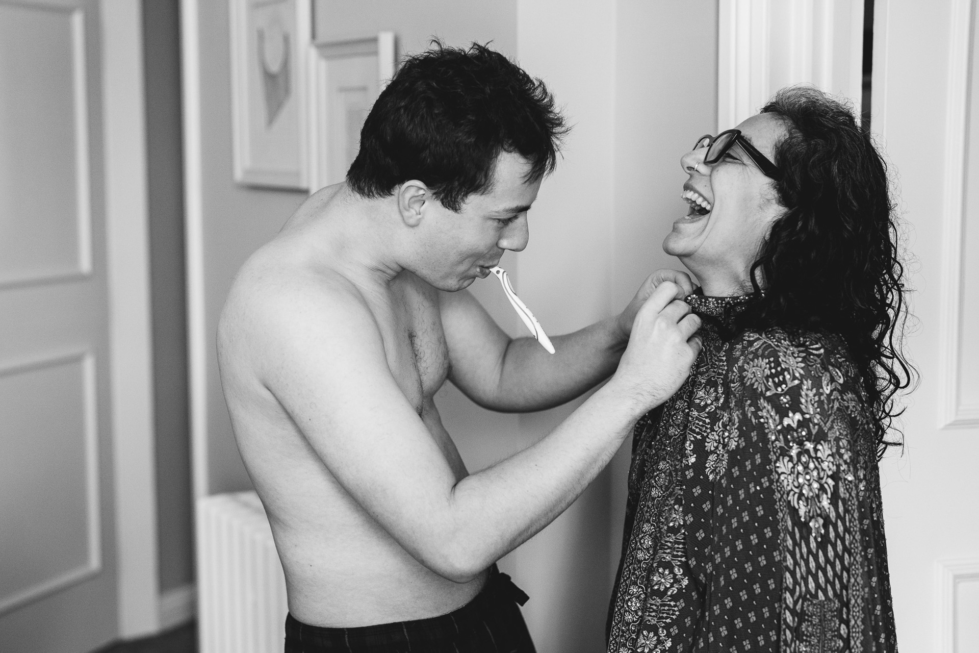 A groom to be, brushing his teeth before his wedding and simultaneously helping his mother with a button as they laugh together