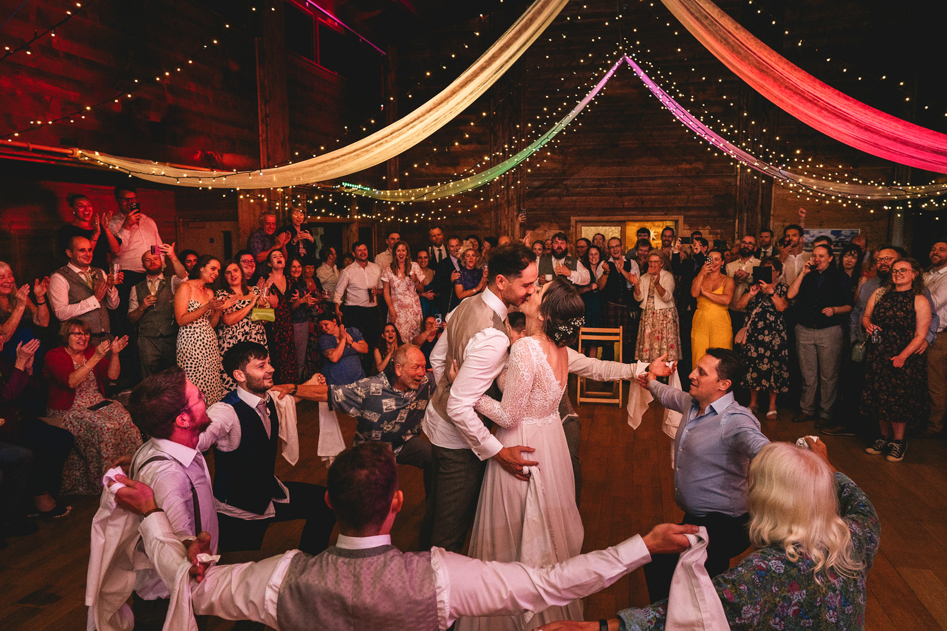 A bride and groom surrounded by morris dancers, kissing in the middle of the dance floor with guests all smiling and clapping around them at a village hall wedding celebration in Devon 