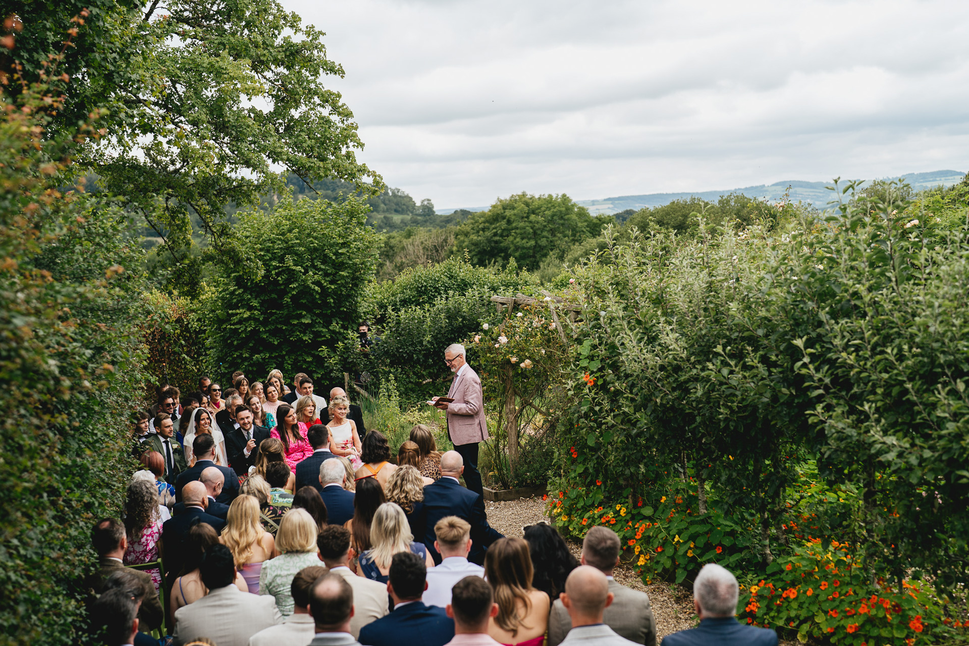 A wide shot of an outdoor wedding ceremony in the kitchen garden at River Cottage, with Devon views behing