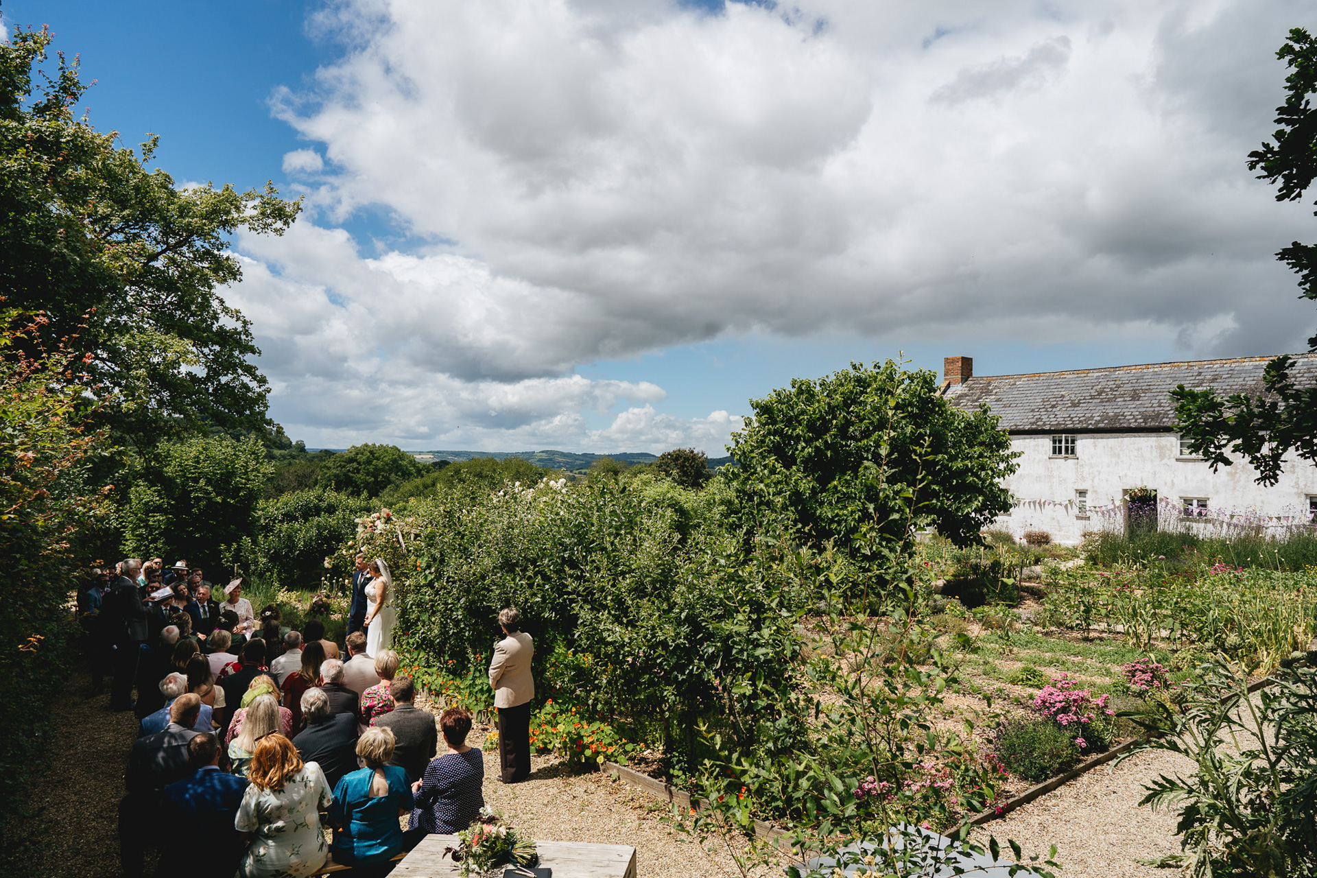 A wide shot of an outdoor wedding ceremony in the kitchen garden at River Cottage, with the farmhouse and Devon views behind