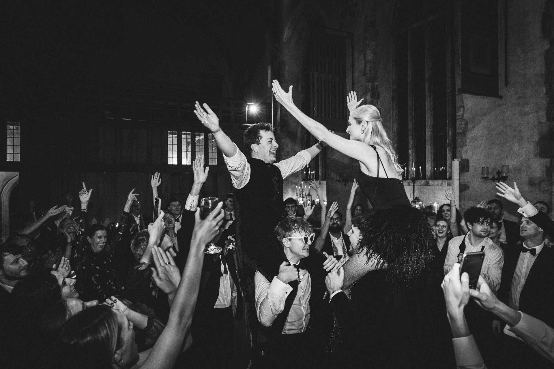 A bride and groom on the dancefloor at Dartington Hall in Devon, carried on the shoulders of their friends and raising their arms in the air in celebration