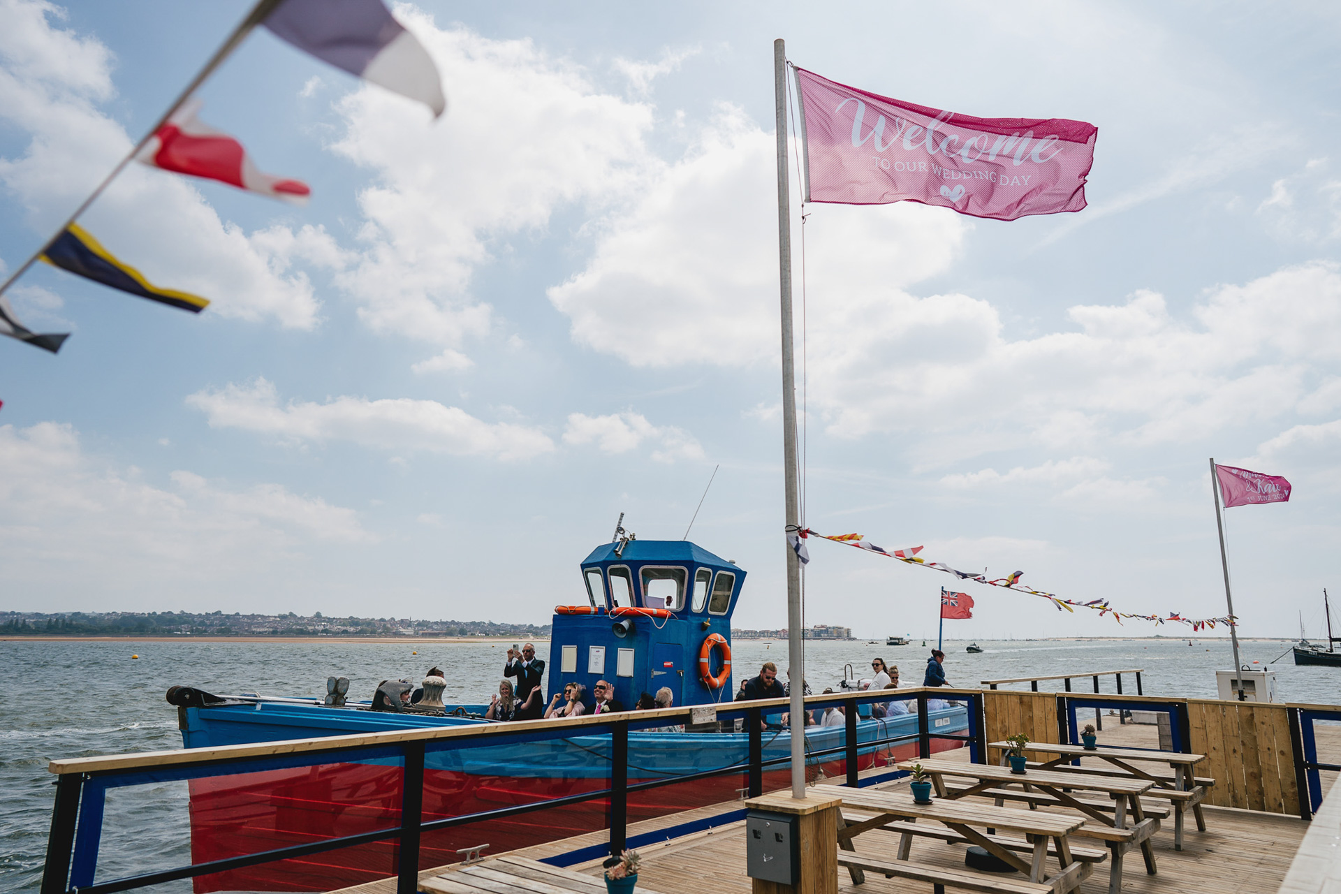 An estuary scene with a flag flying, saying welcome to our wedding day, with a boat arriving behind, filled with wedding guests. 