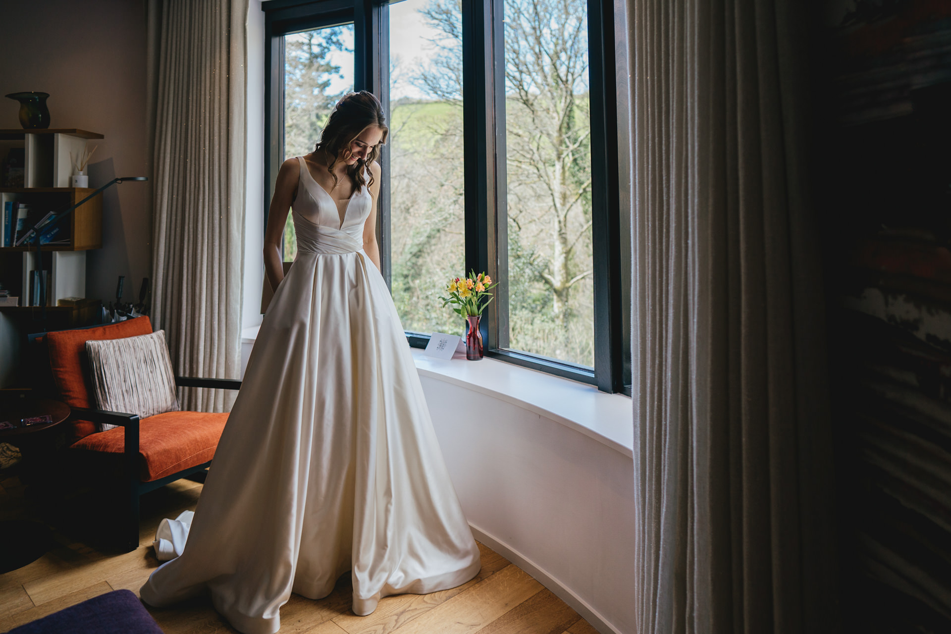 A bride to be in a wedding dress with pockets, standing by a window before her wedding ceremony in Devon