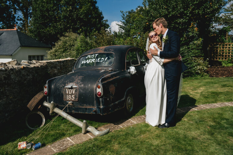 A bride and groom cuddling together by an old car in a family garden at the best Devon wedding