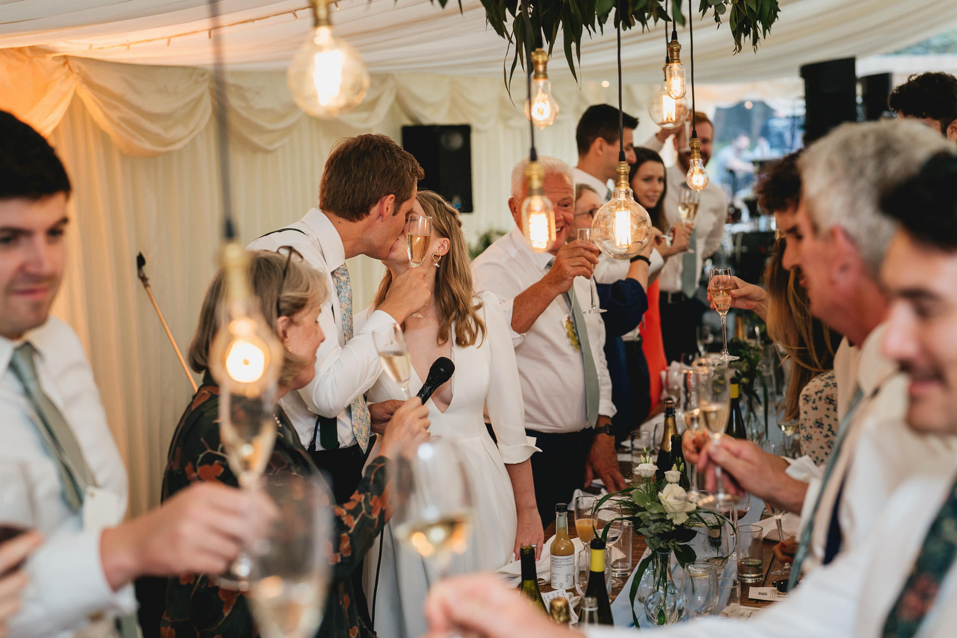 A bride and groom kissing during toasts after speeches at a Devon marquee wedding at home