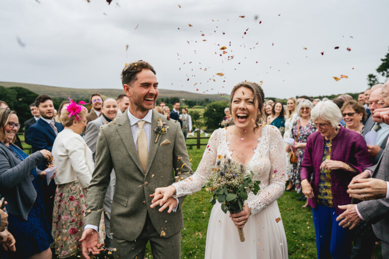A bride and groom walking through confetti and smiling, with Dartmoor behind them.
