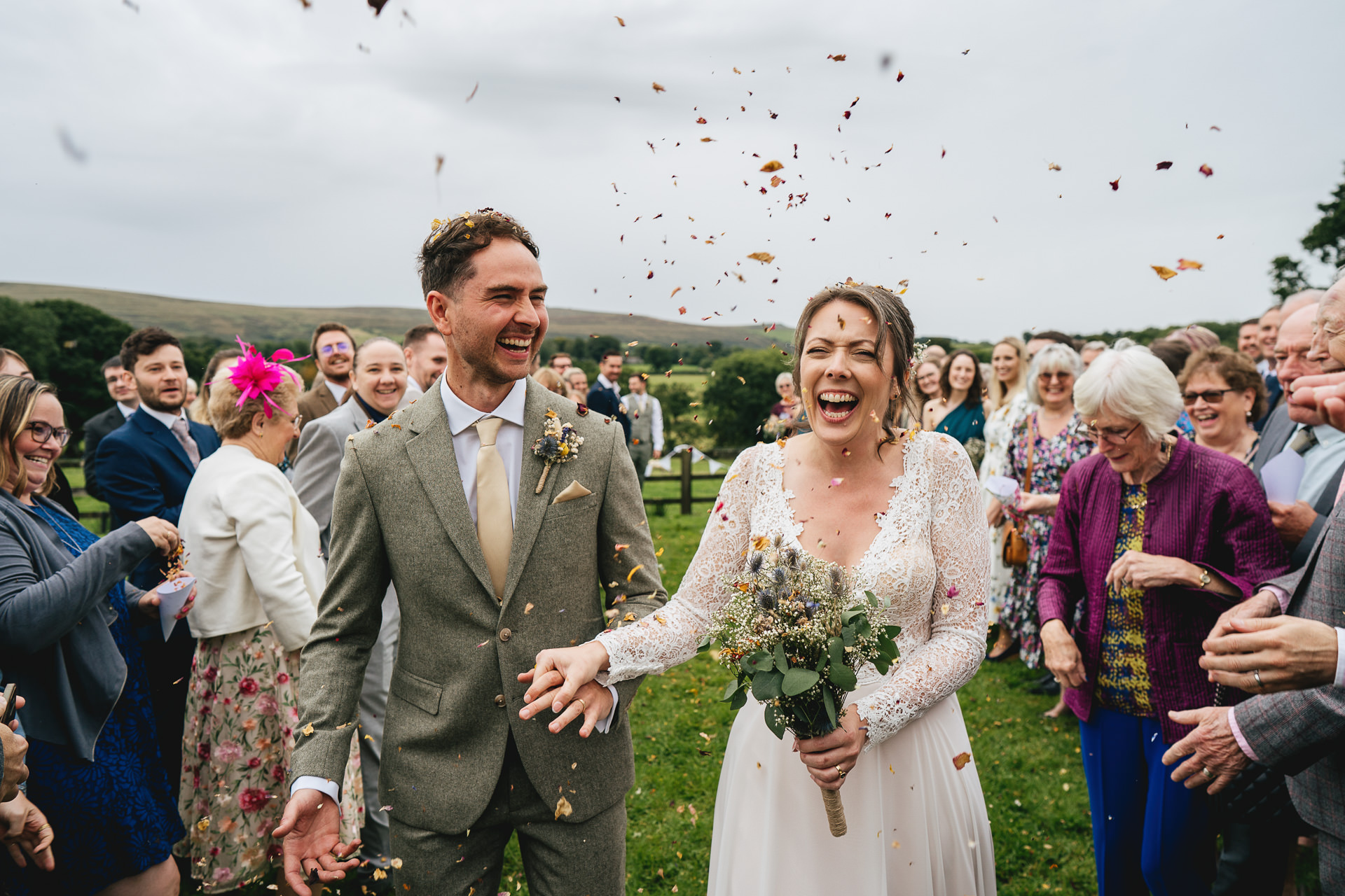 A bride and groom walking through confetti and smiling, with Dartmoor behind them.