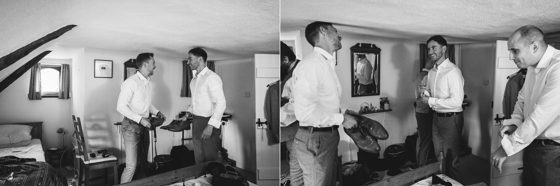 Two men laughing together while getting dressed for a wedding in a cottage bedroom with a low ceiling