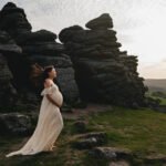 Maternity photo session with a pregnant woman standing by rocks in Devon, wearing a white dress and her hands cradling her bump, with the sun setting over Dartmoor.