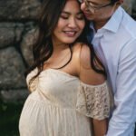 Maternity photo of a pregnant woman standing with a man behind her, cuddling together with their hands on her bump.