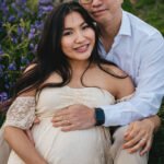A maternity photo outdoors with bluebells behind, with a pregnant woman sitting down, with a man sitting behind her, cuddling together with their hands on her bump.