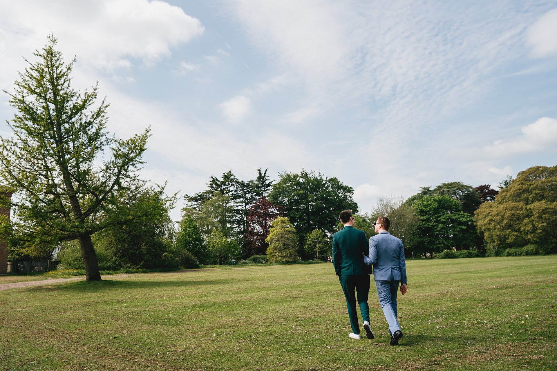 Grooms walking arm in arm across a grass field with trees in the background after their wedding ceremony