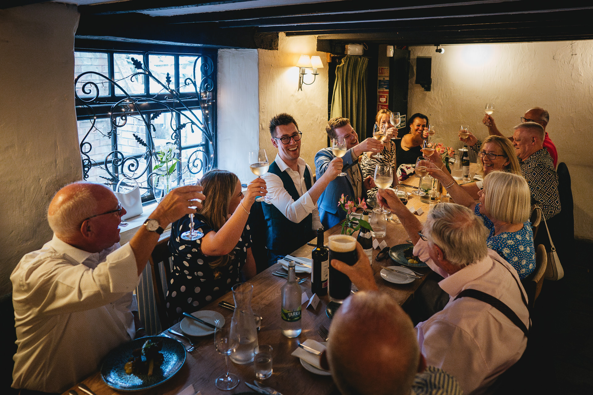 A small group celebrating an elopement wedding, seated around a long table at Rendezvous Wine Bar in Exeter. The group are all raising glasses to say cheers.