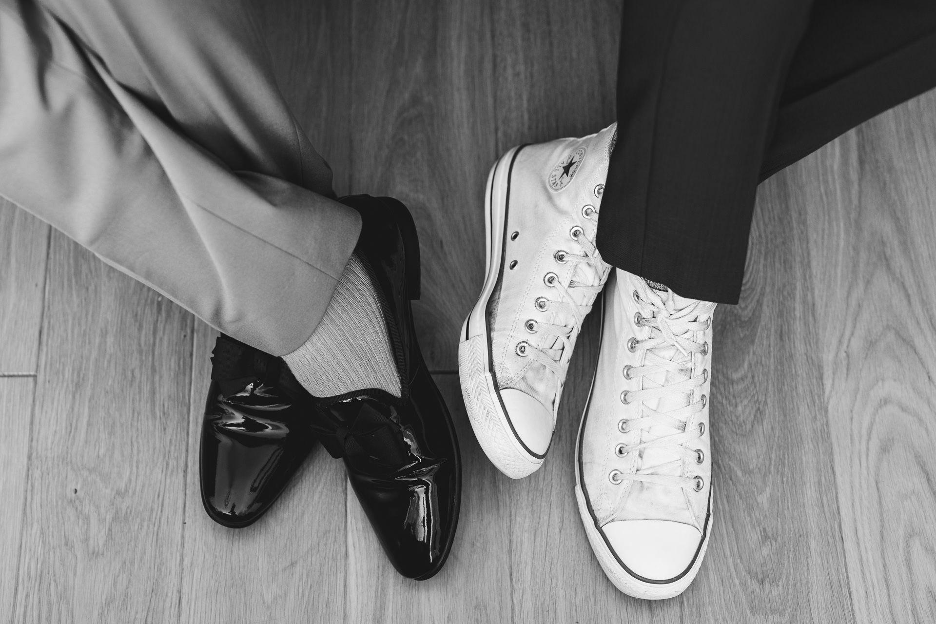 A black & white image of the feet of the grooms, with smart black shoes on one side and white converse high tops on the other