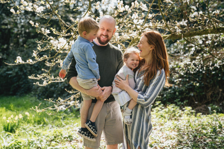 A family photography session with parents holding children and laughing together under a magnolia in full bloom at an arboretum near Exeter in Devon