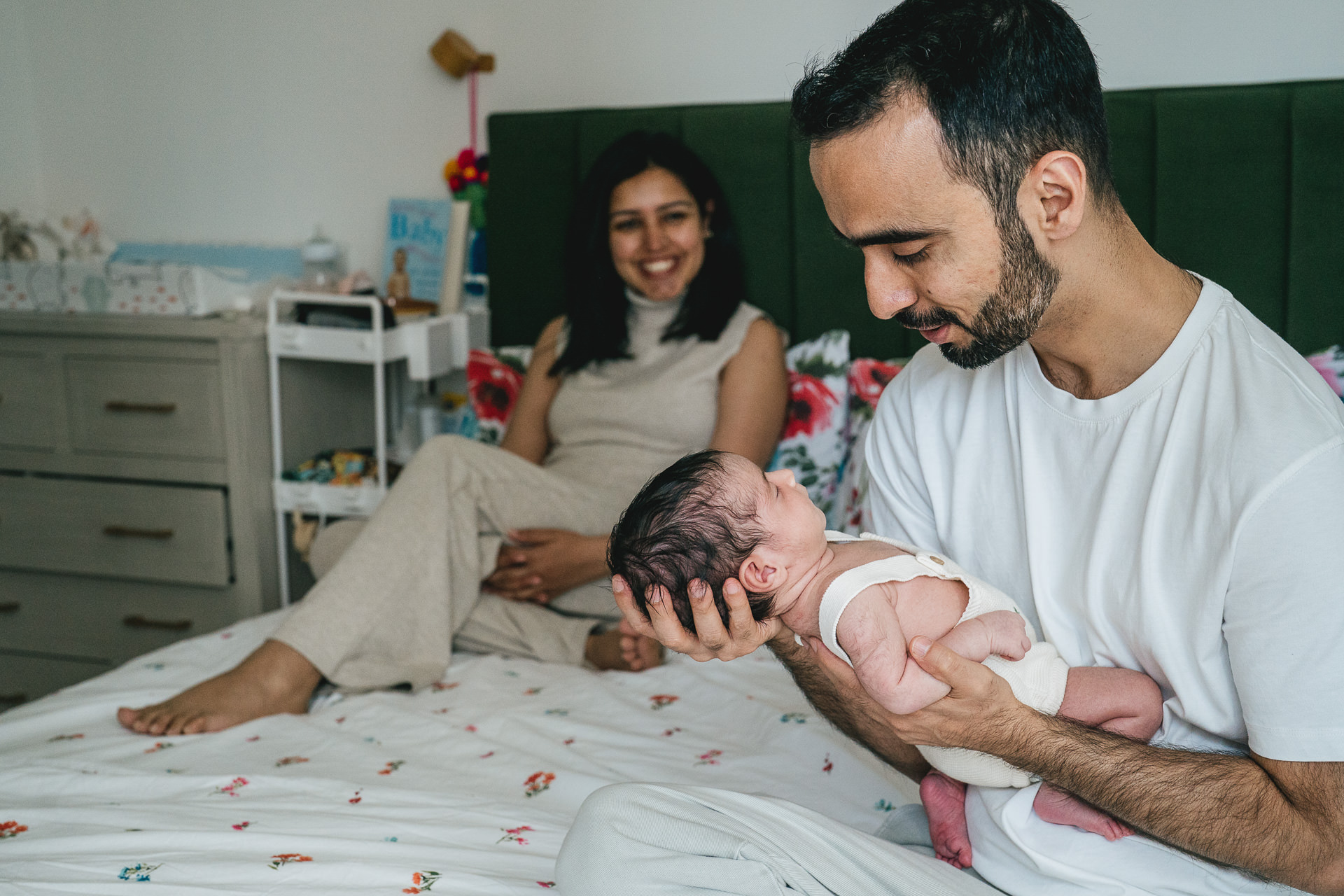 New parents sitting on a bed together and smiling at a newborn baby