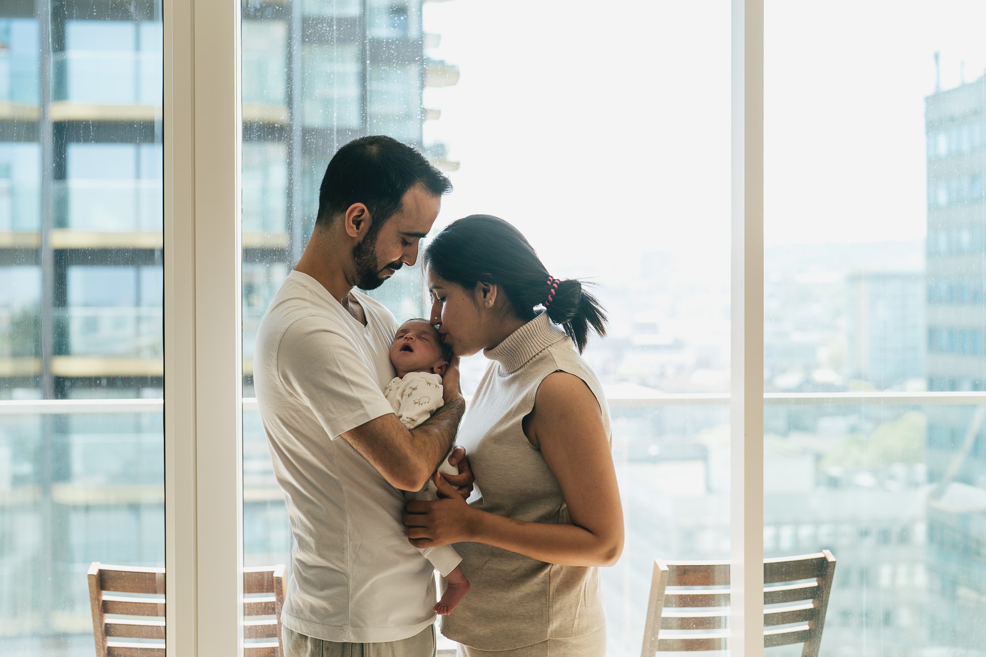 A newborn baby photography session at home in London, with parents standing together by a large window and holding their baby