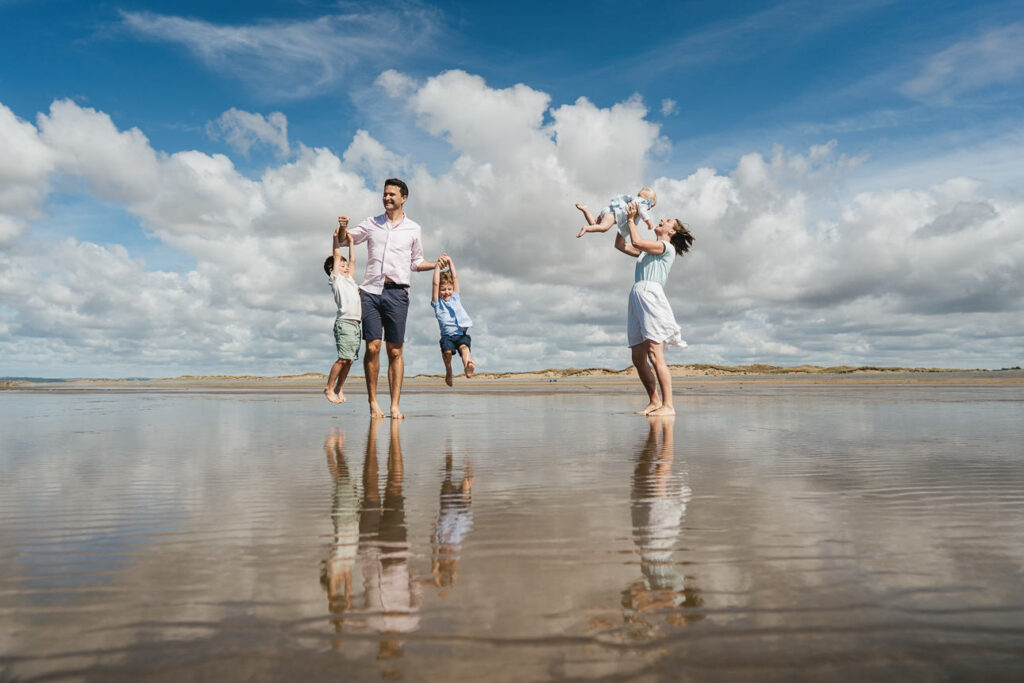 A family group during a photography session on the beach together, with two boys hanging off their father's arms and mother holding a baby in the air