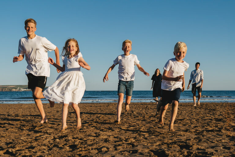 A family photography session on the beach in Devon with four children running towards the camera smiling and two parents in the background laughing