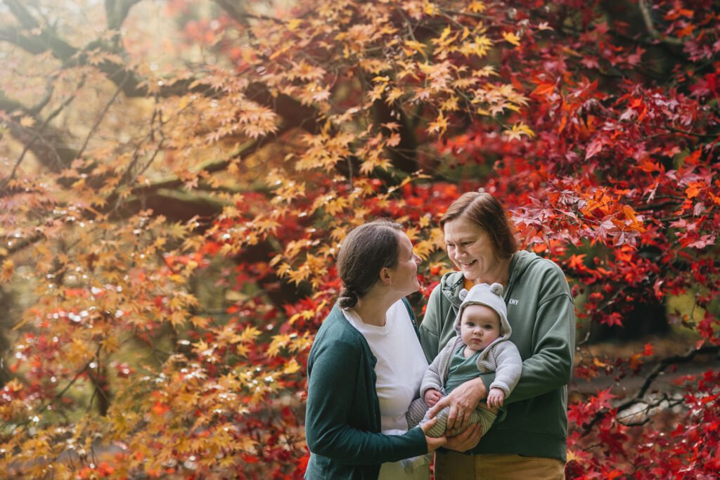 Two mothers with their young baby, smiling at each other with red and orange autumn leaves behind them.