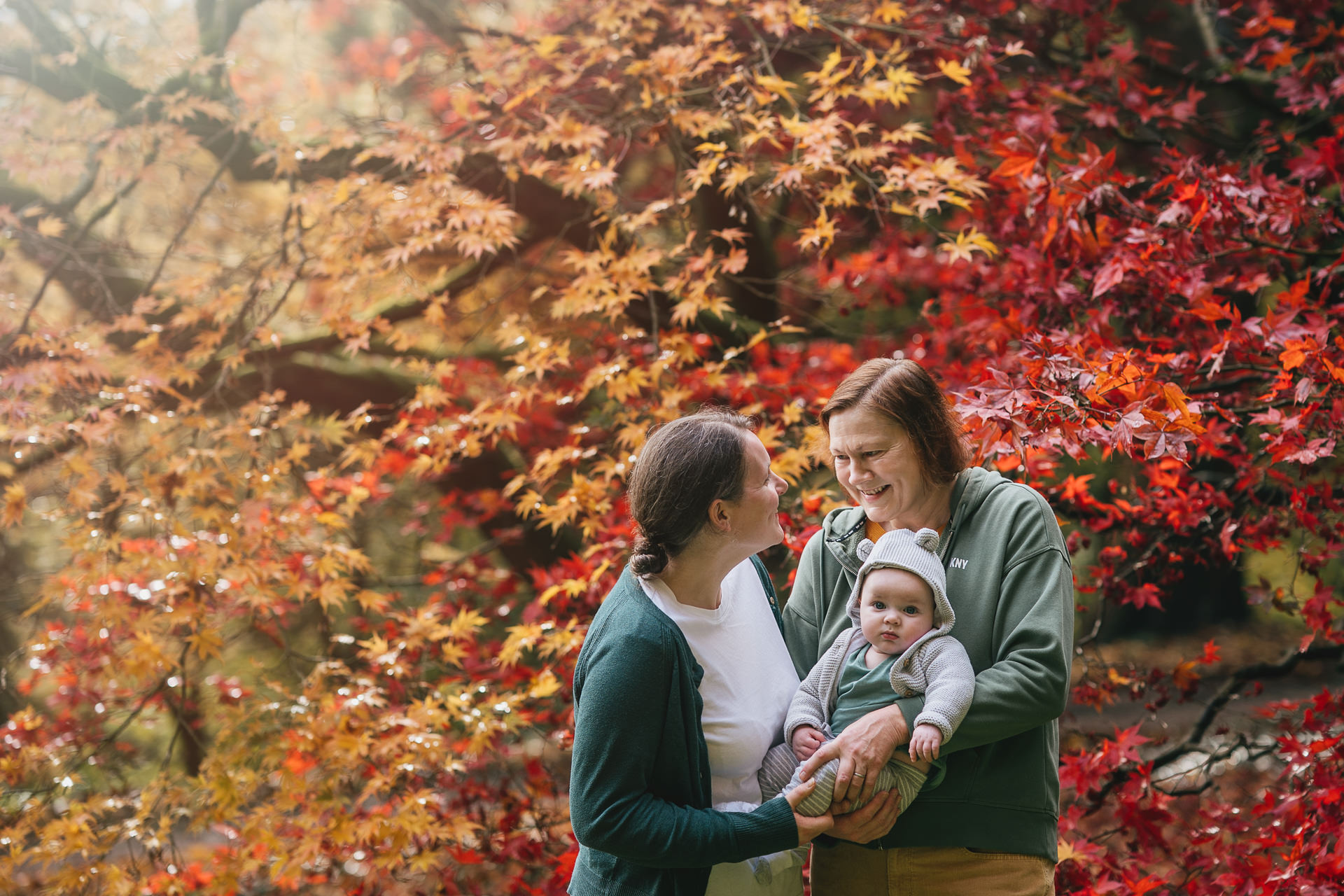 Two mothers with their young baby, smiling at each other with red and orange autumn leaves behind them. 