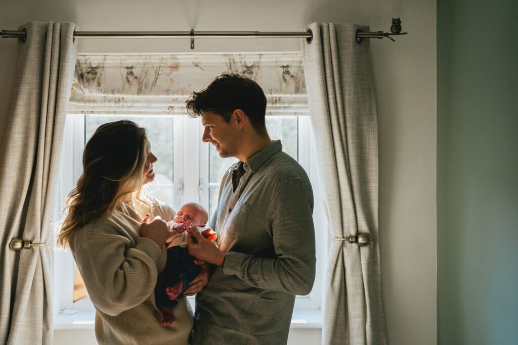 Parents standing by a window together, holding a newborn baby boy during a family photography session in Devon