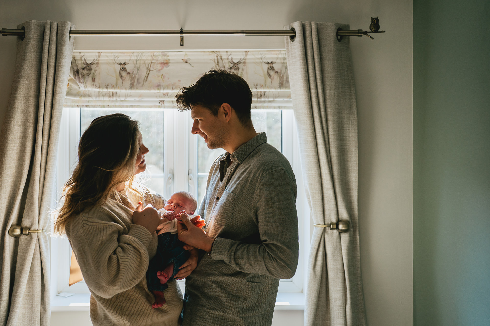 Parents standing by a window together, holding a newborn baby boy during a family photography session in Devon