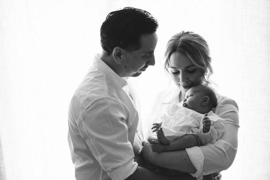 A mother and father standing by a window, cradling their newborn baby.