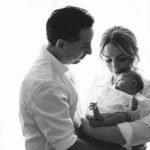 A mother and father standing by a window, cradling their newborn baby.