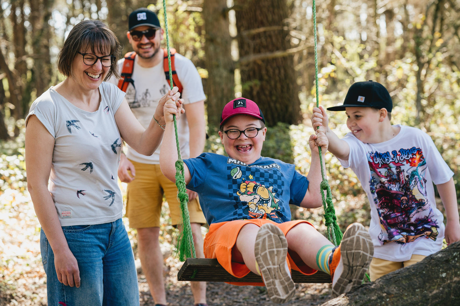 A relaxed family portrait with two boys playing on a swing together in woodland, and mother and father laughing with them. 