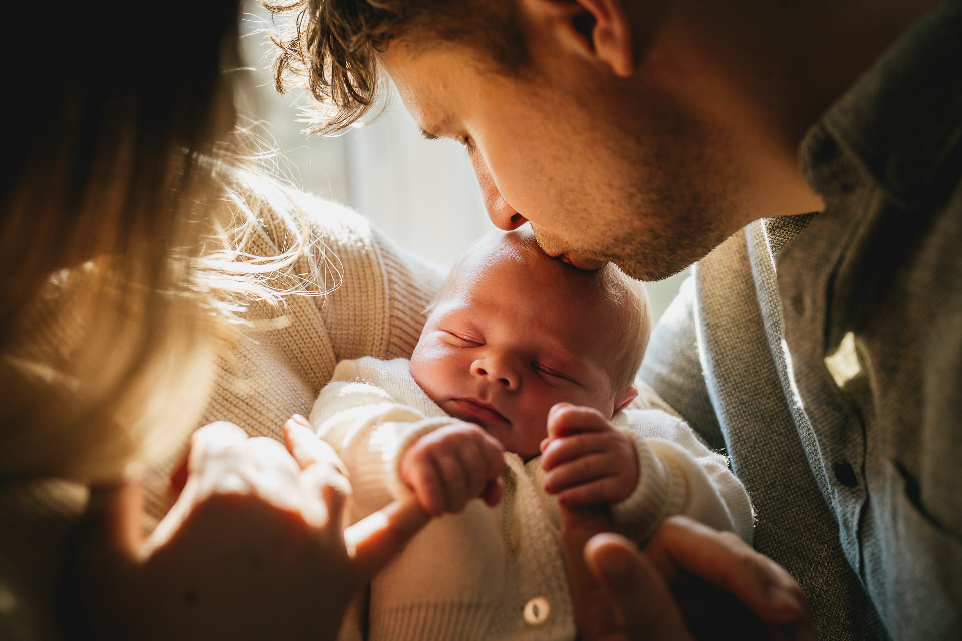 A close up photo of a mother and father cradling a newborn baby with the father kissing the baby's head. .