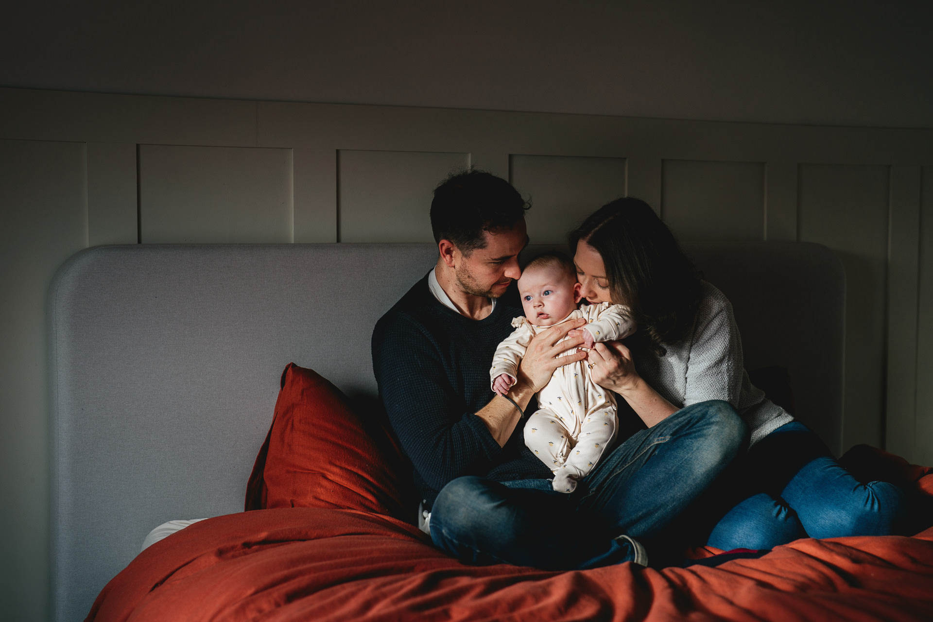 A couple sitting together on a bed with a red cover, cuddling a new baby, in a patch of window light. 