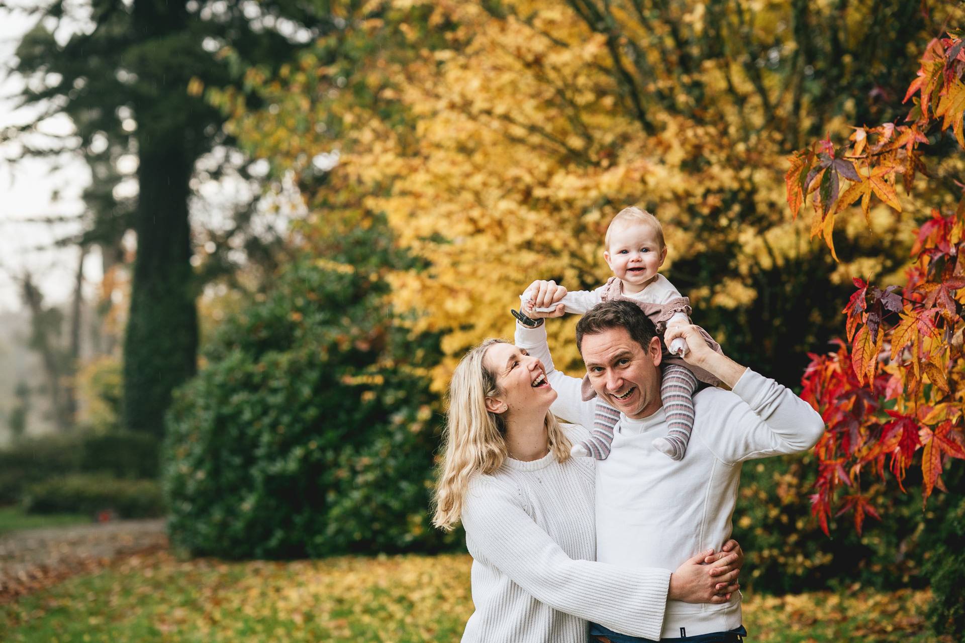 Two parents with a young baby, laughing together with autumn leaves on the trees behind them. 