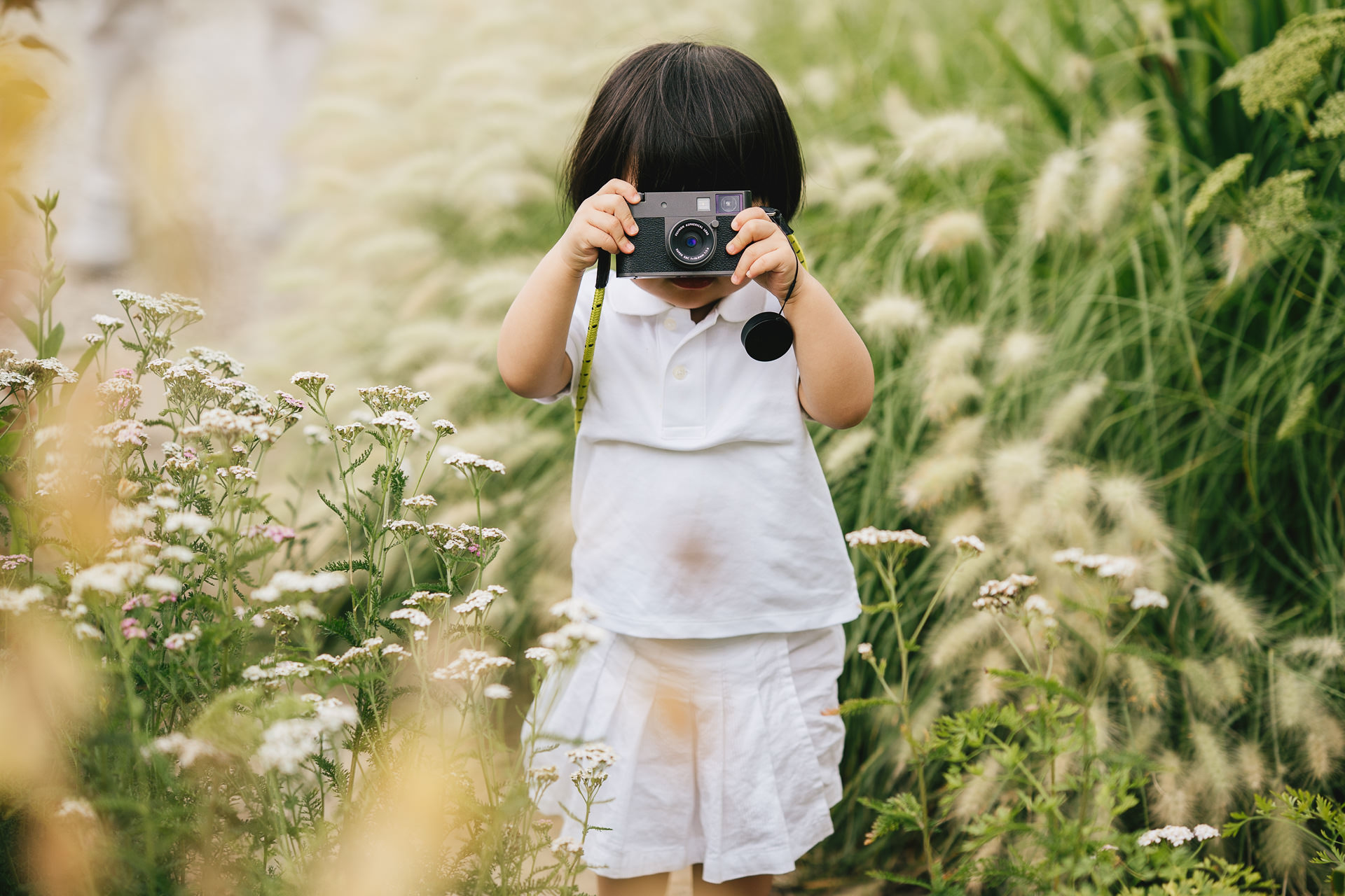 A young toddler dressed in white, holding a camera to her face and taking a photo of the photographer during a family photography session at the Newt in Somerset