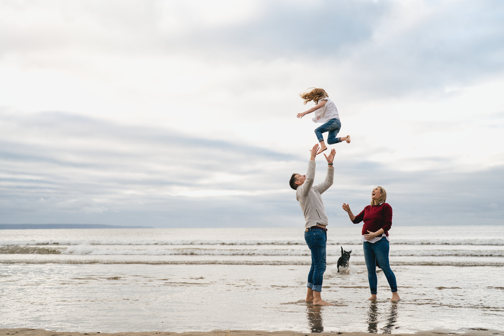 A family playing together during a photography session at Saunton Sands, with a pregnant mother laughing and watching a father throw a young girl in the air. 