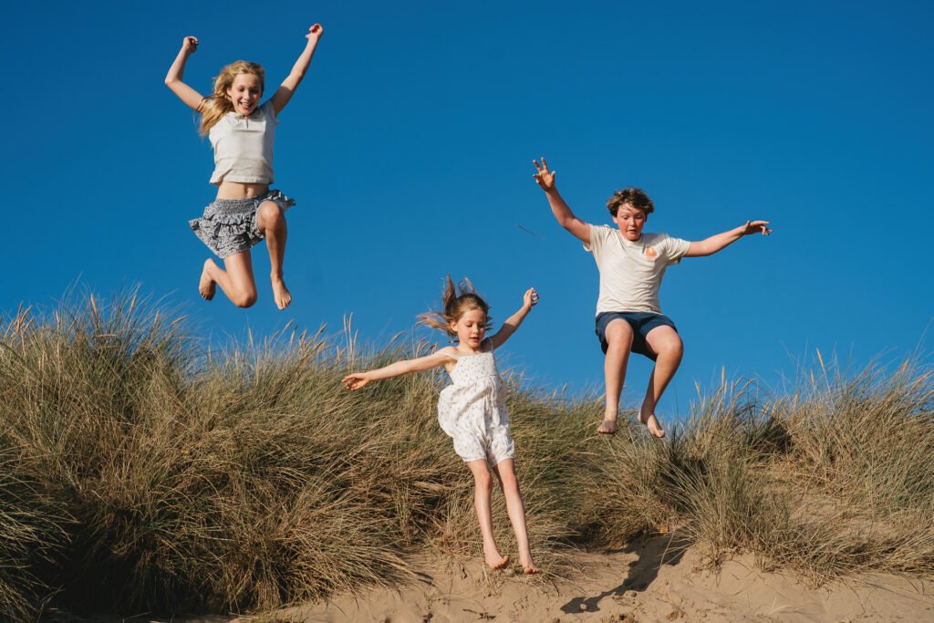 Teenage children during a family photography session in South Devon, jumping off a sand dune with blue sky above