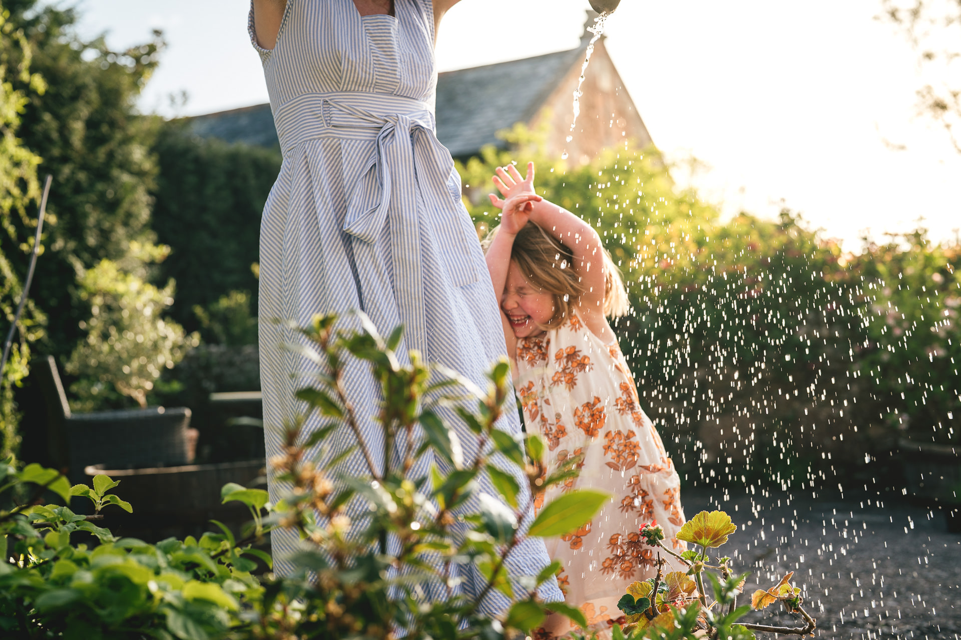 A young girl laughing and sheltering from water as she plays with her mother, watering plants at home during a family photography session in Devon
