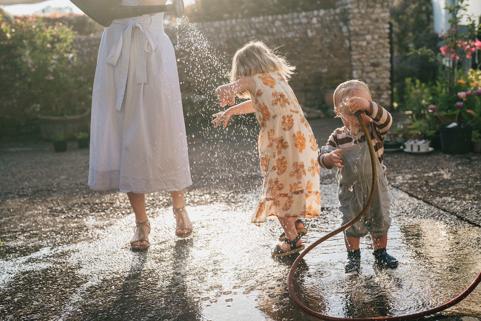 Two young children playing with a watering can and hose during a family photography session at home in Devon in the evening sunlight