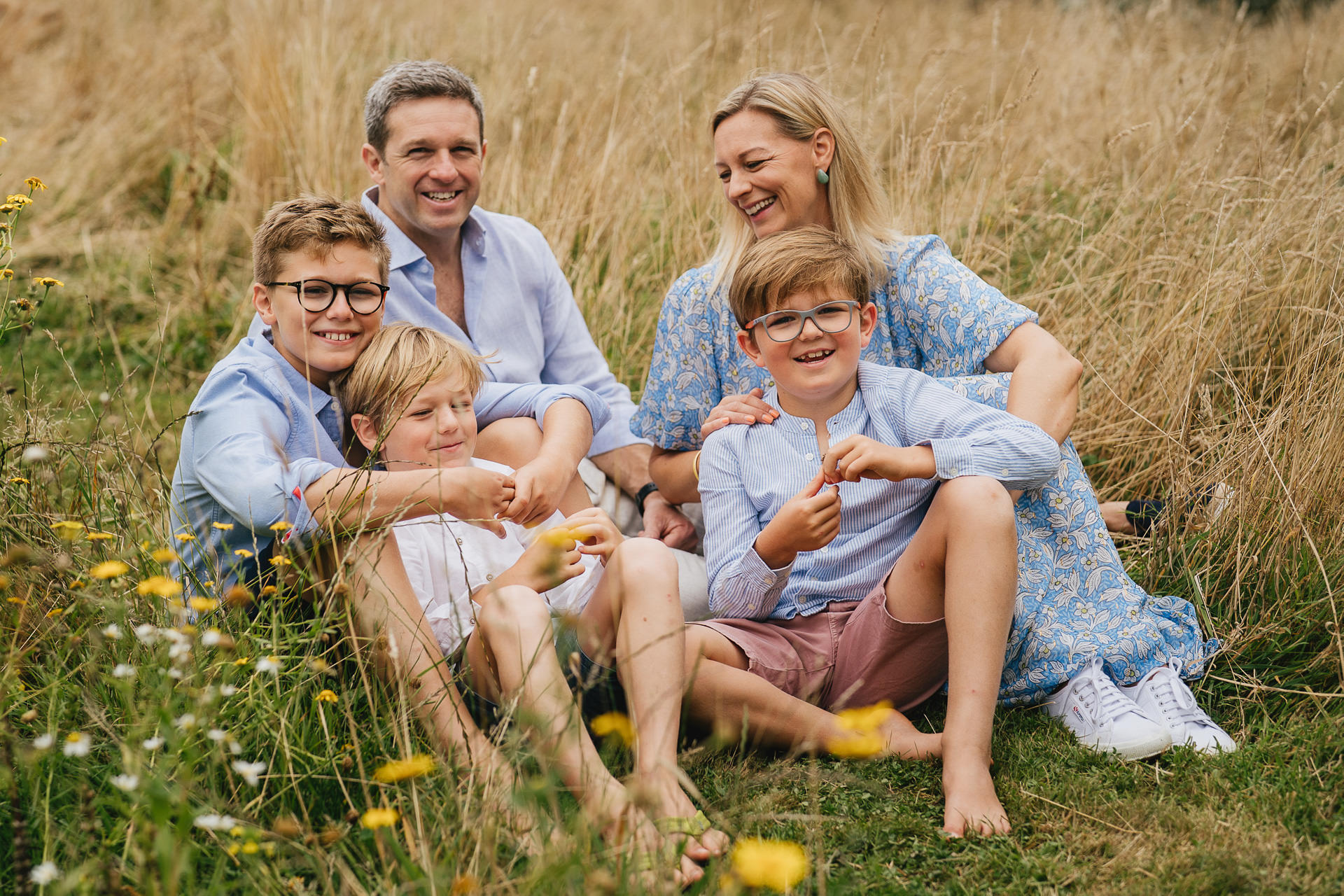 A relaxed family group photo from an extended family photography session in Devon with parents sitting amongst long grass with three boys