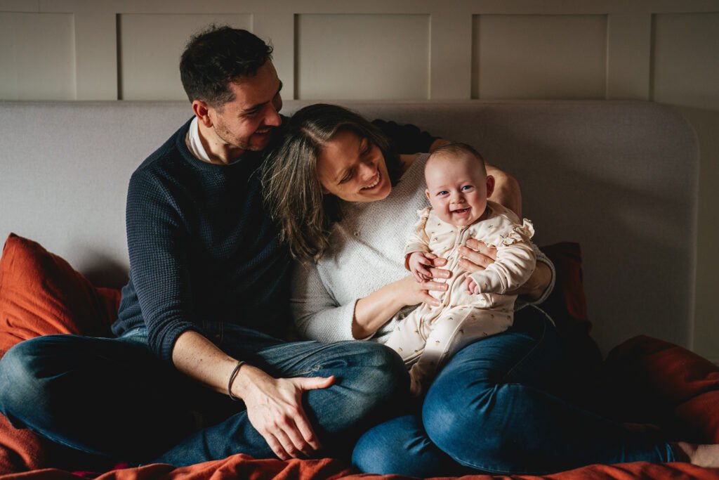 Parents with a smiling baby girl, sitting on their bed together in North Devon