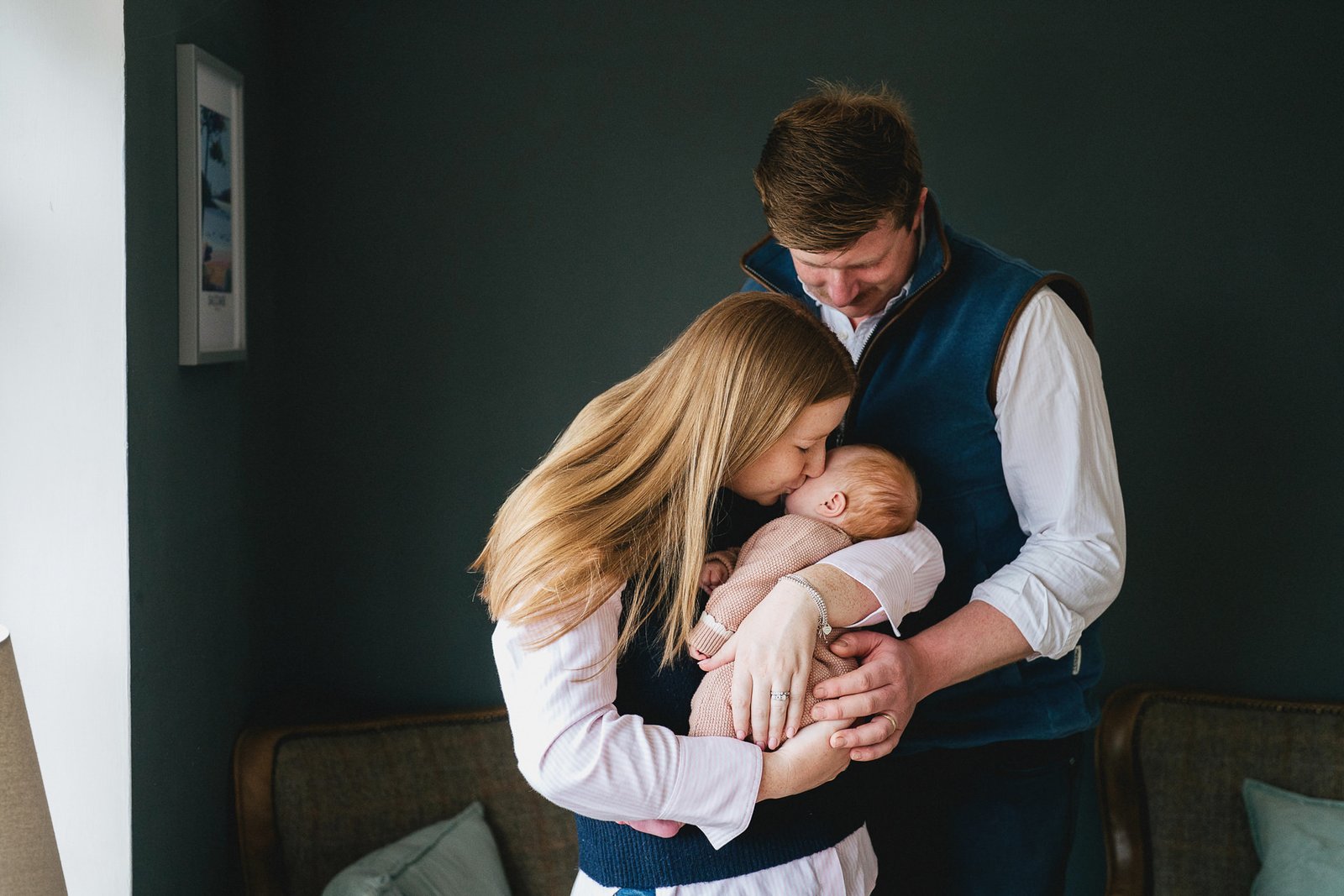 Parents cuddling a new baby at home for a baby photo session with a dark blue wall behind,