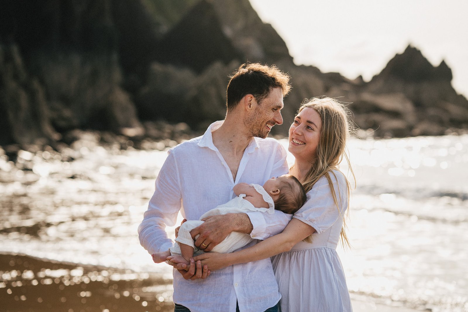 Parents cuddling a young baby and smiling at each other during a photography session on the beach 